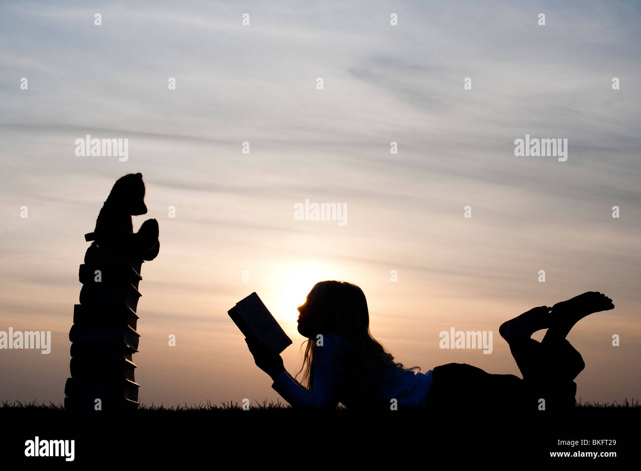 Ragazza giovane disteso la lettura di un libro di fronte a un orsacchiotto seduto su una pila di libri al tramonto. Silhouette Foto Stock