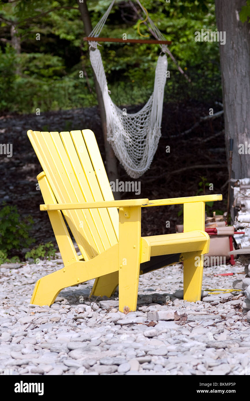 Un lone giallo sedia Adirondack su una spiaggia di pietra, con amaca in background. Foto Stock