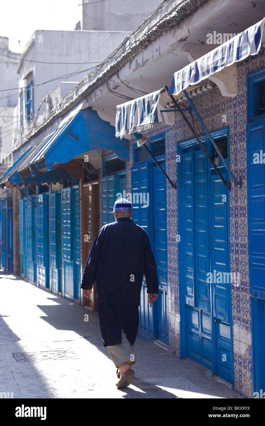 Uomo che indossa una calotta passeggiate lungo una strada lastricata che in passato il blu cobalto con persiane chiuse bancarelle del nord della Medina di Essaouira Foto Stock