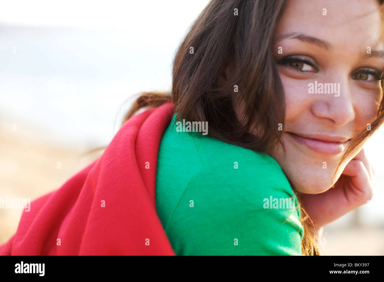 Femmina nel suo inizio 20's avvolto in una coperta rossa sorrisi come guarda la fotocamera sulla spiaggia. Foto Stock