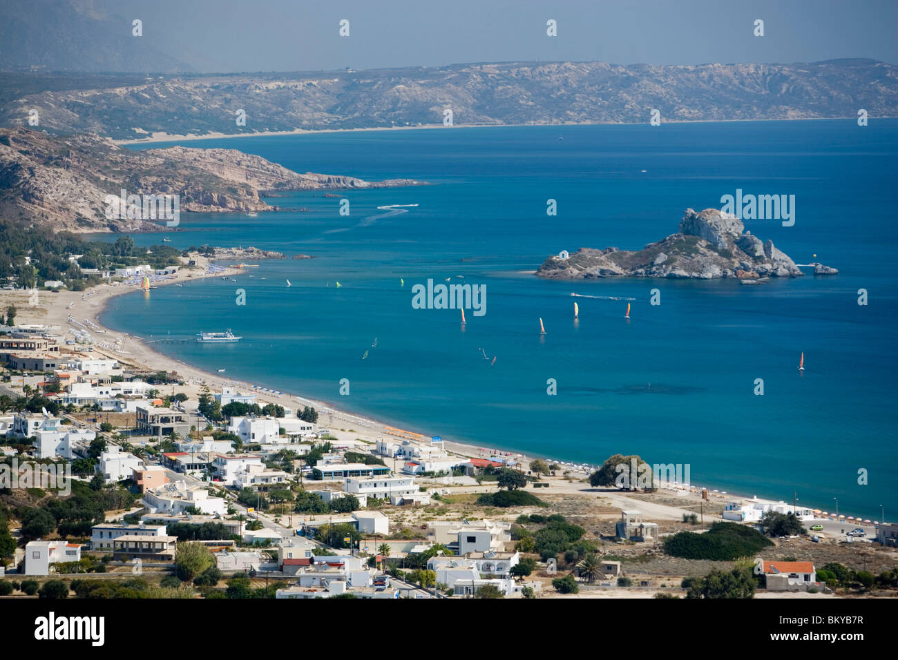 Vista sulla spiaggia e sulla baia di Kefalos con surfer di Kastri isola con la cappella di San Nicolò, Kefalos, Kos, Grecia Foto Stock