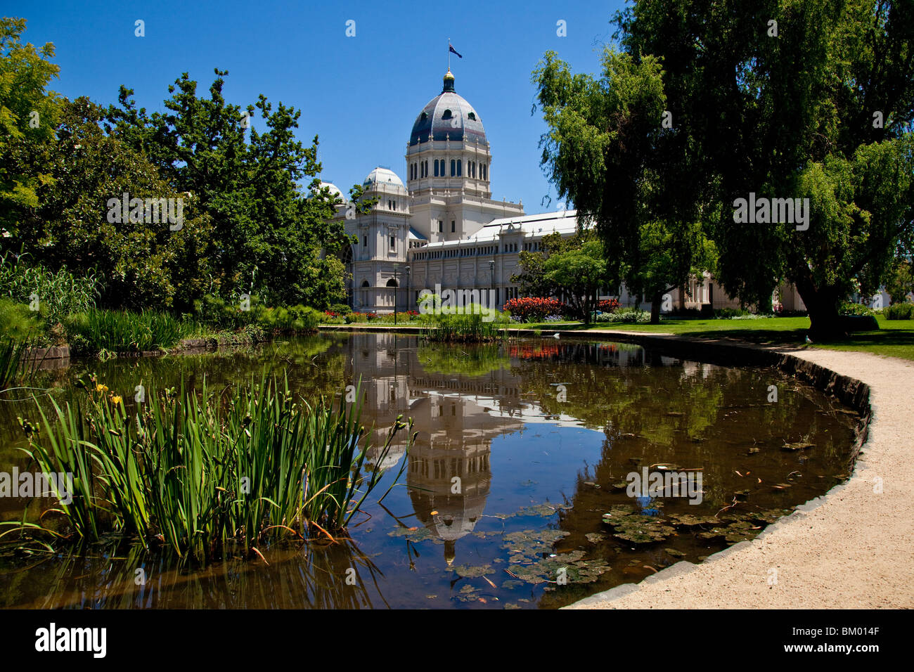 Royal Exhibition Building, Melbourne e giardini Carlton si riflette in un stagno affiancato da un percorso e alberi maturi al sole. Foto Stock