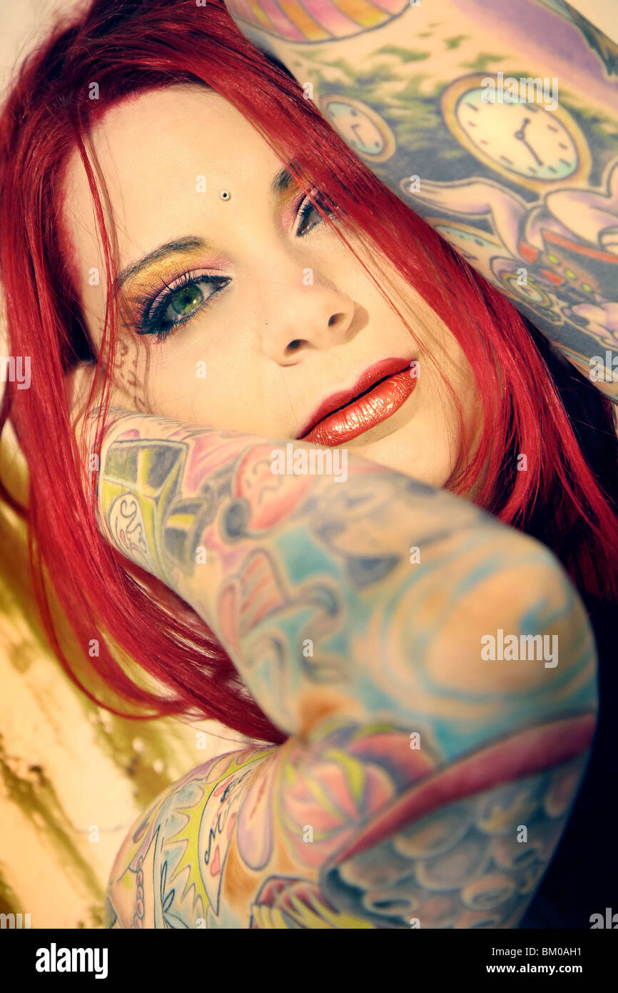 Primo piano di una donna di colore rosso per i capelli e gli occhi verdi con le braccia coperte di tatuaggi colorati Foto Stock