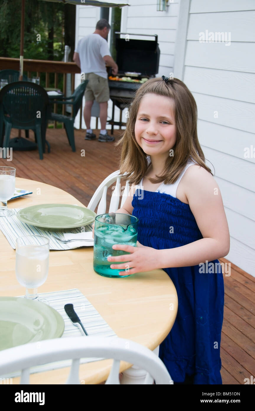 Una giovane ragazza imposta la tabella mentre suo padre le grigliate alimenti sul barbecue in preparazione per un partito all'aperto. Foto Stock