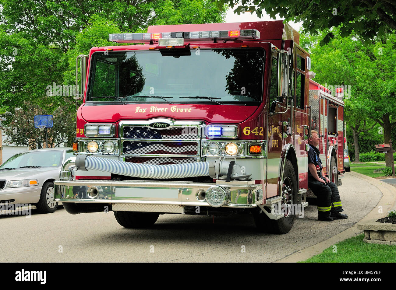 Vigile del fuoco in appoggio sul fuoco motore durante una chiamata di emergenza in una casa suburbana. Nota patriottica bandiera americana graphic sui carrelli grill. Foto Stock