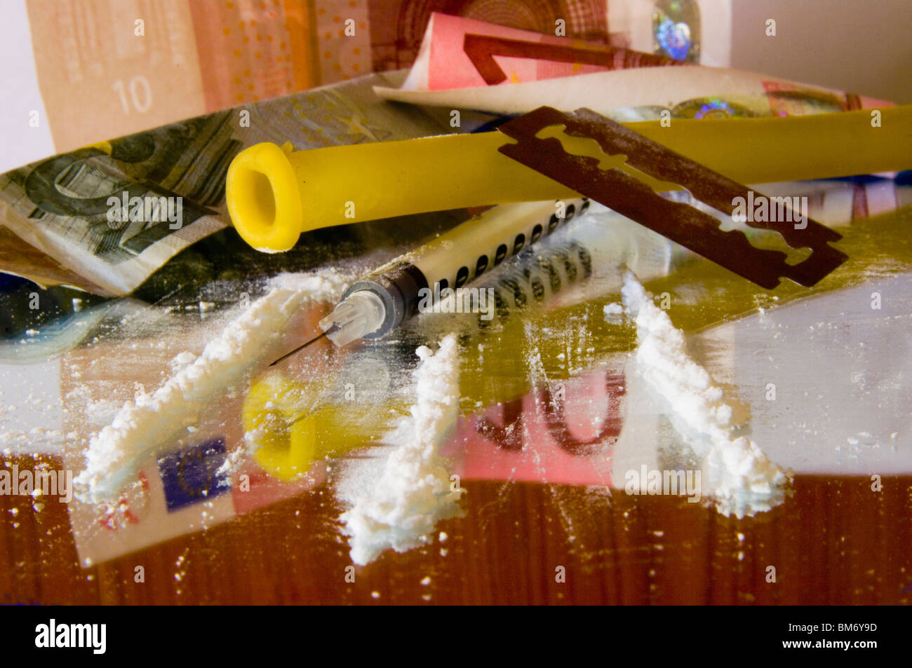 Close-up mostra piste di cocaina che convergono verso una siringa di insulina un laccio emostatico la lama di un rasoio con sfondo euro Foto Stock