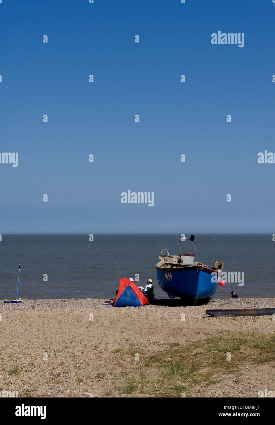 Vacanzieri rifugiandosi dietro una barca da pesca sulla spiaggia di Sizewell nel Suffolk. Foto di Gordon Scammell Foto Stock