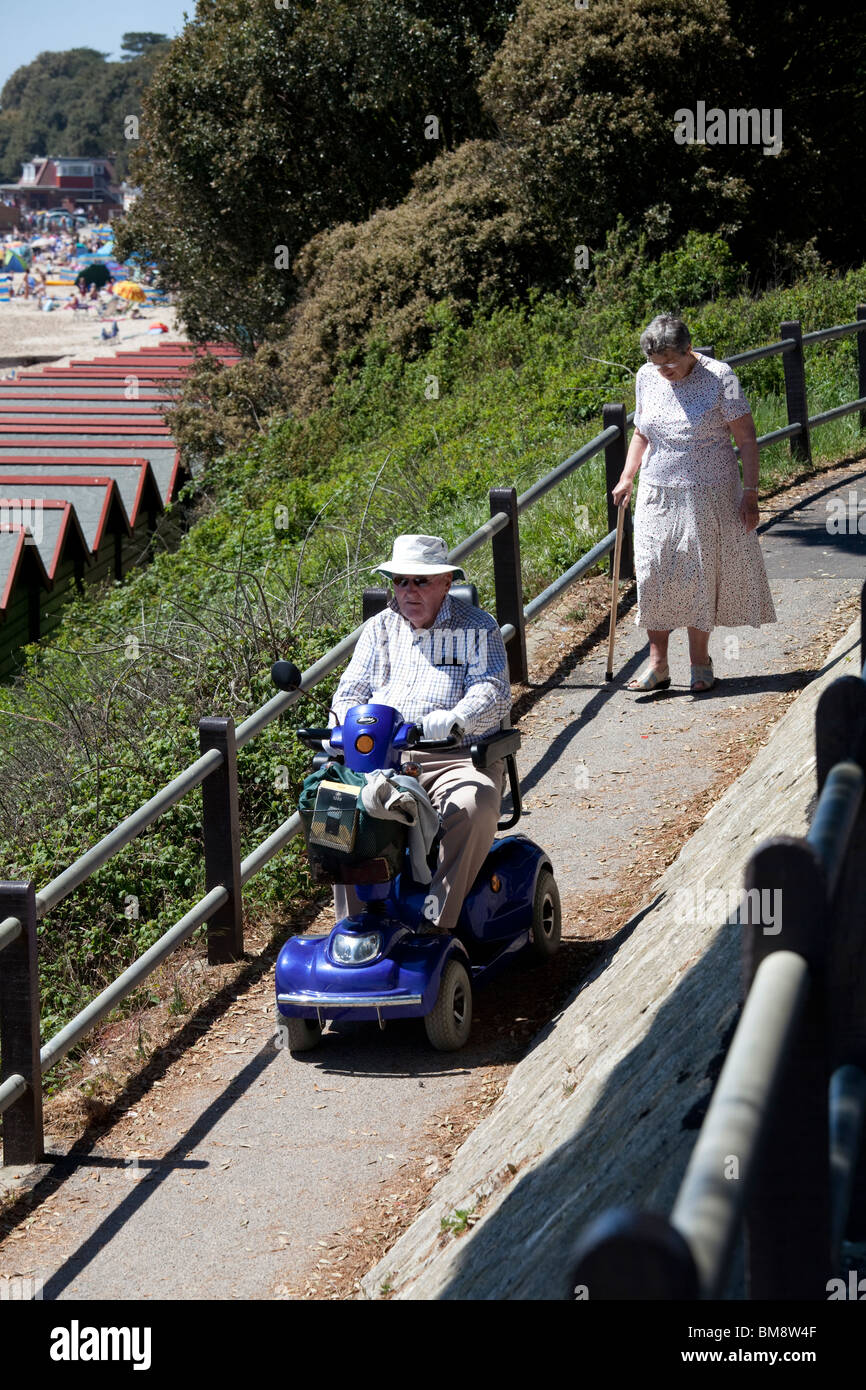 Il vecchio gentleman nel cappello per il sole sui disabili motorizzata buggy seguita da una vasta con bastone scendendo la rampa verso la spiaggia Foto Stock