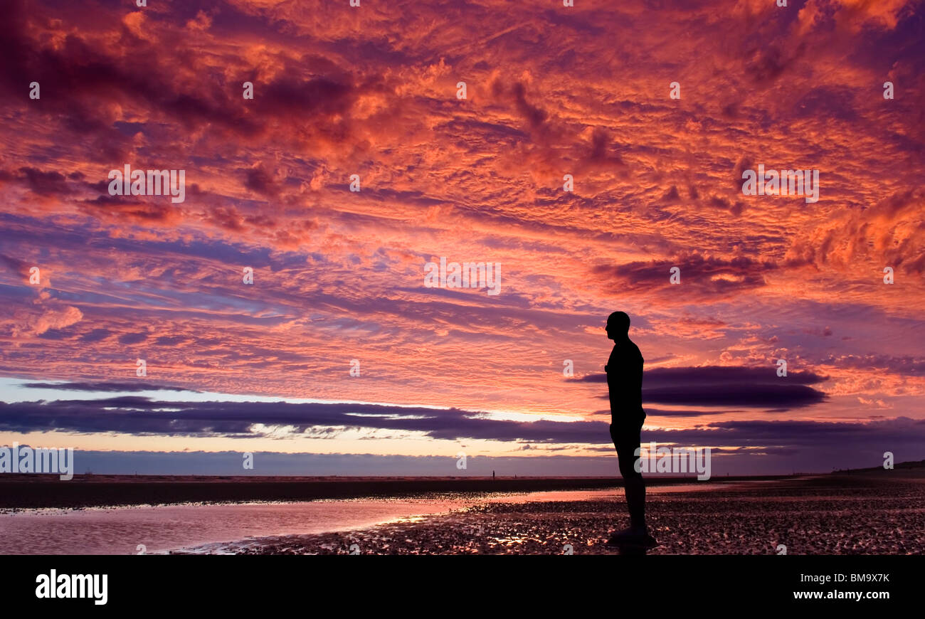Antony Gormleys 'UN ALTRO POSTO' Crosby Beach Merseyside. Singola "l'uomo del ferro" stagliano contro un drammatico tramonto rosso Foto Stock