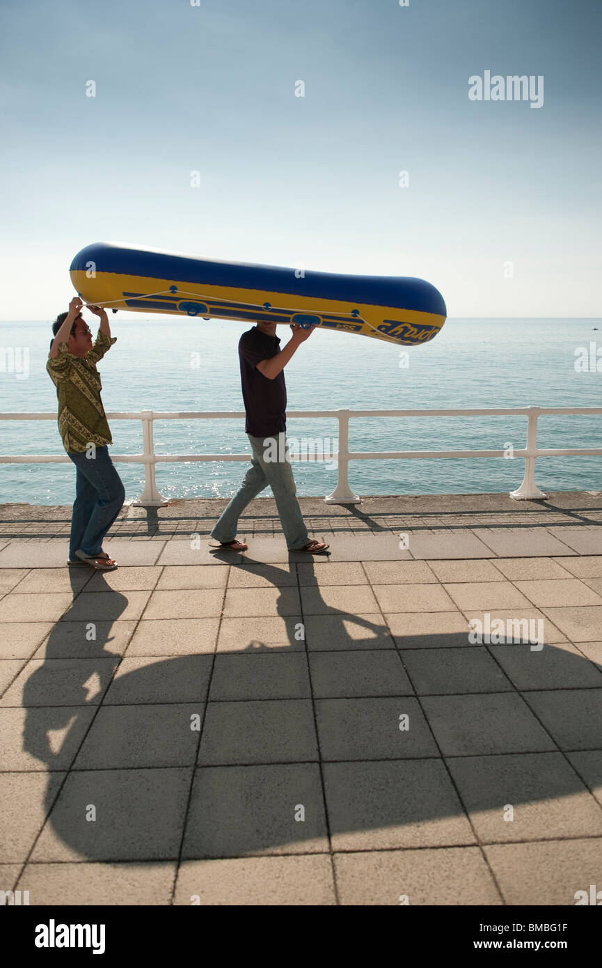 Due giovani studenti che trasporta un canotto gonfiato a Aberystwyth Beach, Cardigan Bay, West Wales, pomeriggio estivo, REGNO UNITO Foto Stock