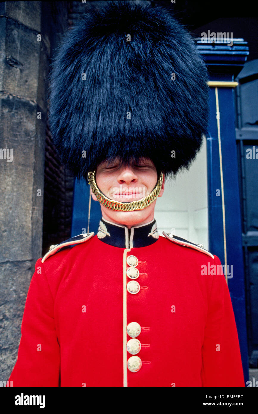 Un schivo British palace guard nel suo tradizionale abito rosso tunica e bearskin black hat cerca di nascondere un sorriso mentre sul dovere di Londra, Inghilterra. Foto Stock