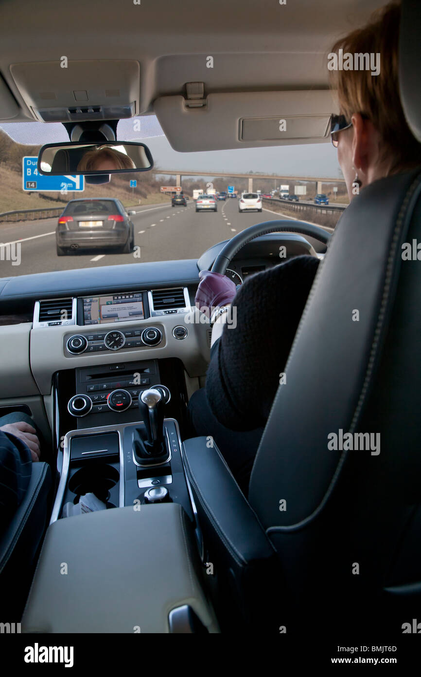Interno del Range Rover Auto essendo pilotato da donna in autostrada in UK, uomo passeggero, con sat nav sul cruscotto e cartello autostradale Foto Stock