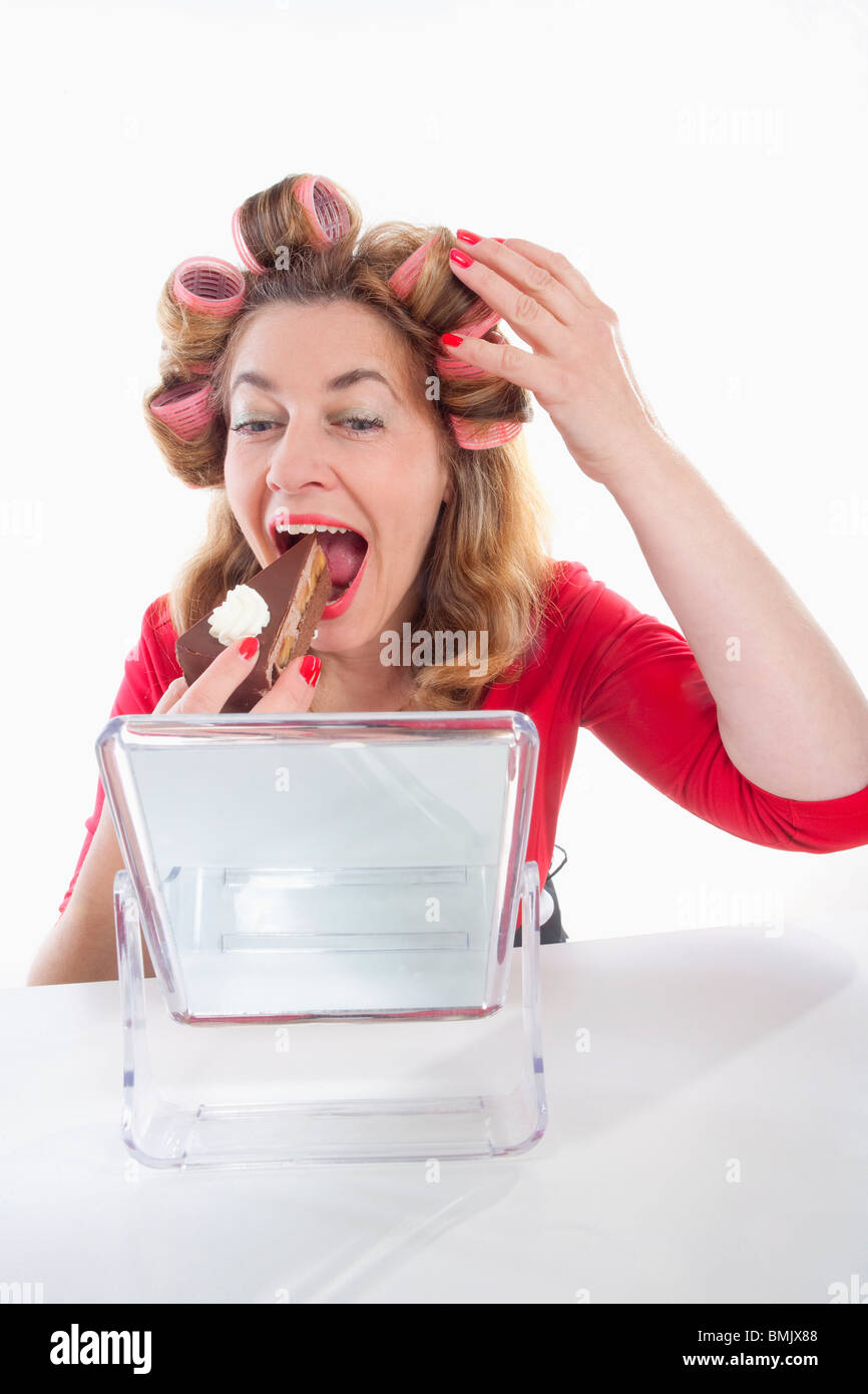 Donna di mezza età con i capelli rulli di mangiare la torta guardando lo specchio Foto Stock
