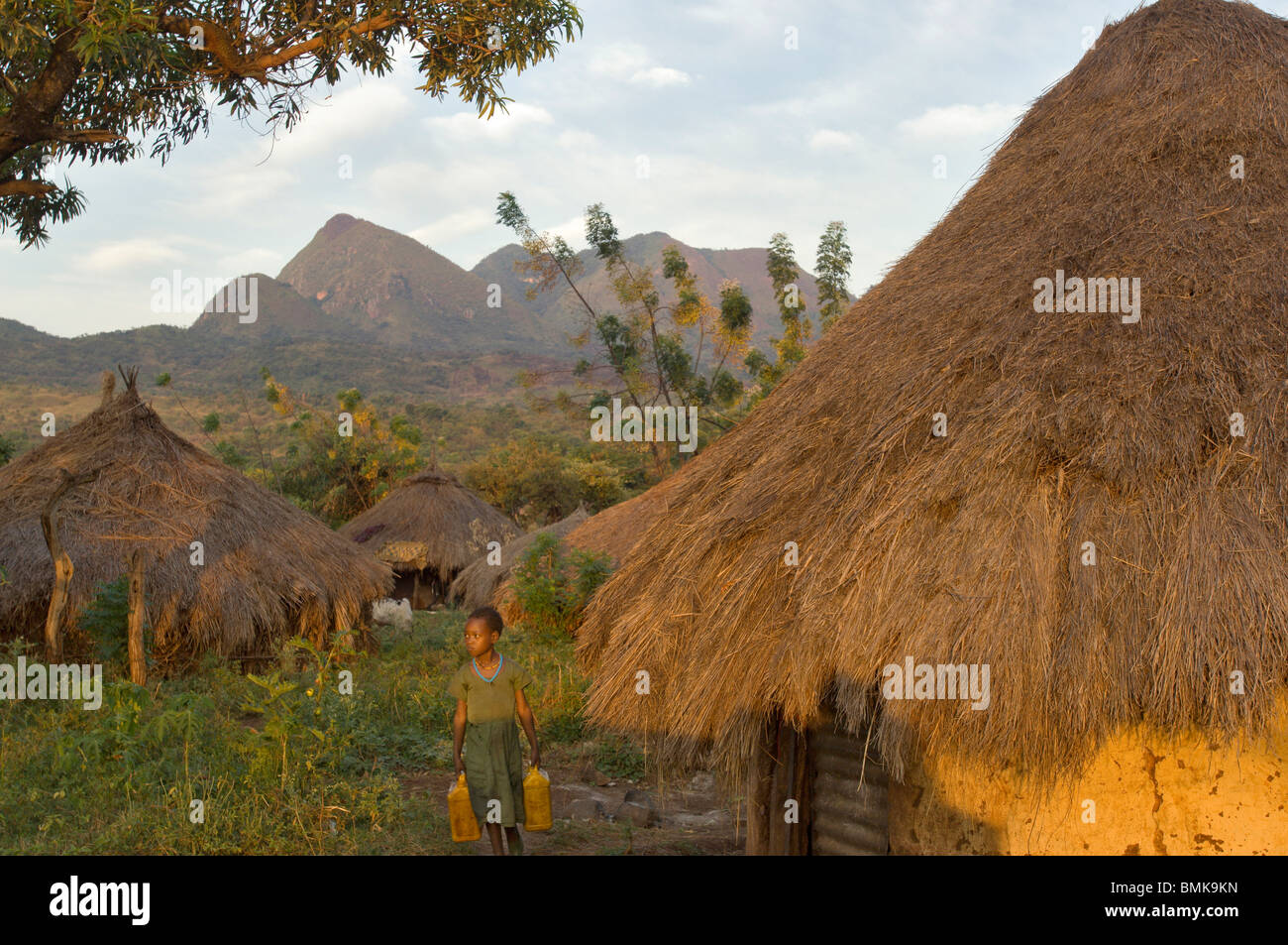 Etiopia, regione dell'Omo, Tulgit. Giovane ragazza in abito verde che trasportano contenitori di acqua e passato la sterpaglia-case con tetti di suri (Surma Tribù). Foto Stock