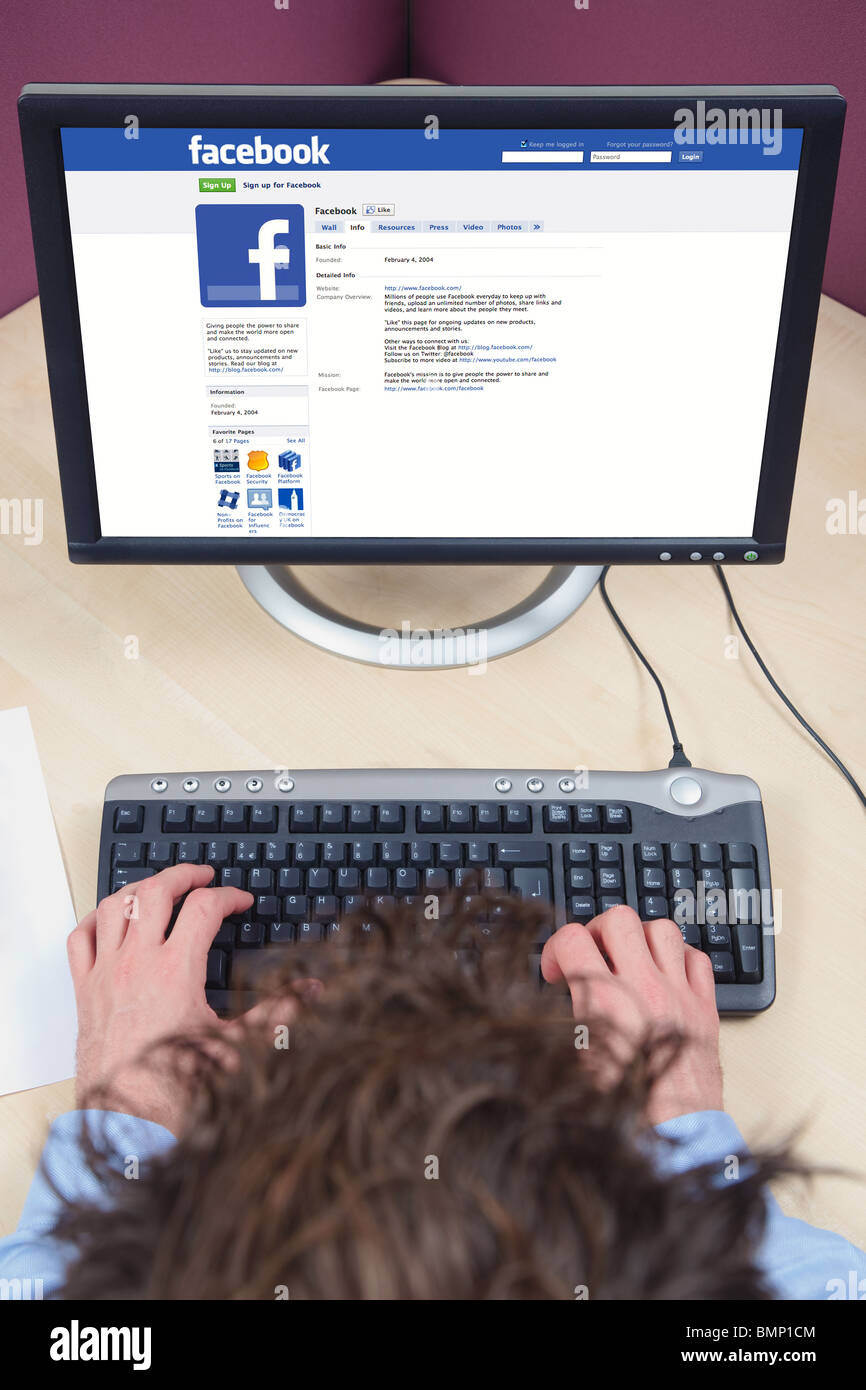 Tettuccio di colpo di un uomo seduto a una scrivania che lavora su un computer, guardando una pagina di Facebook sul suo schermo. Foto Stock