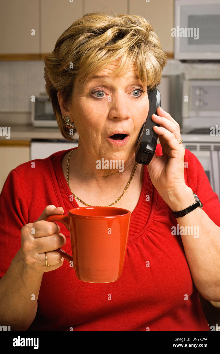 Senior donna, tenendo un rosso tazza da caffè, parla al telefono con uno sguardo di shock e sorpresa sul suo viso. Foto Stock