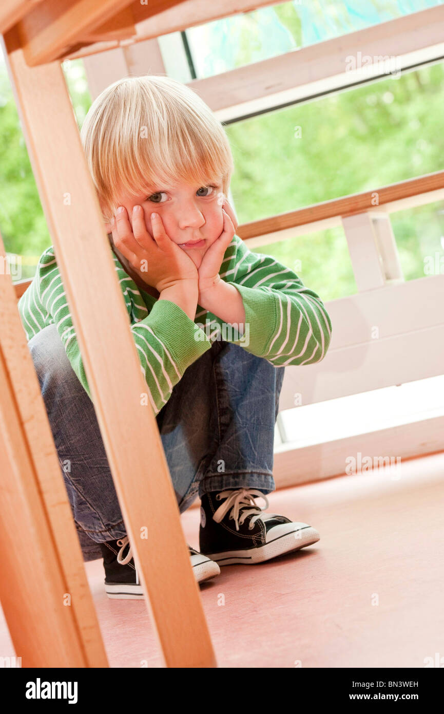 Ragazzo giovane accovacciata accanto a un tavolo e tenendo la testa in mani, basso angolo di visione Foto Stock