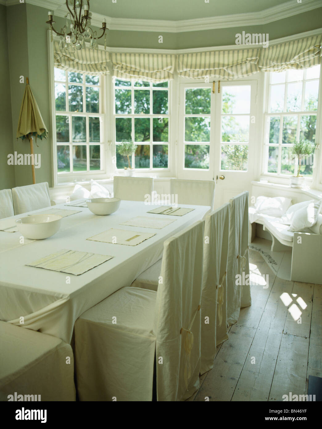 White slip-copre su sedie in crema paese sala da pranzo con un panno di lino sul tavolo di fronte a baia finestra con tende a strisce Foto Stock