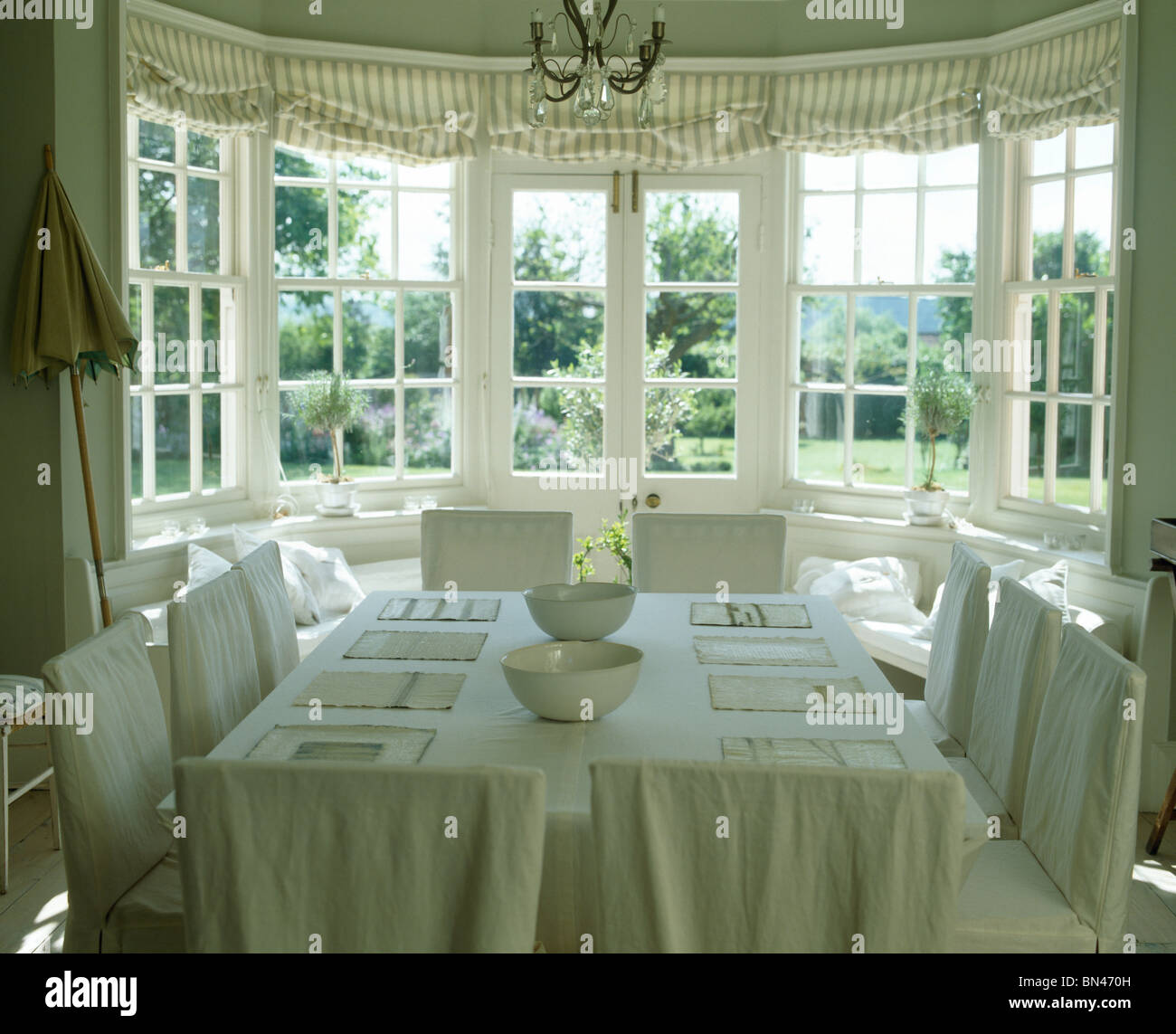 La crema slip-copre su sedie in paese sala da pranzo con tovaglie di lino sul tavolo di fronte a baia finestra con tende a strisce Foto Stock