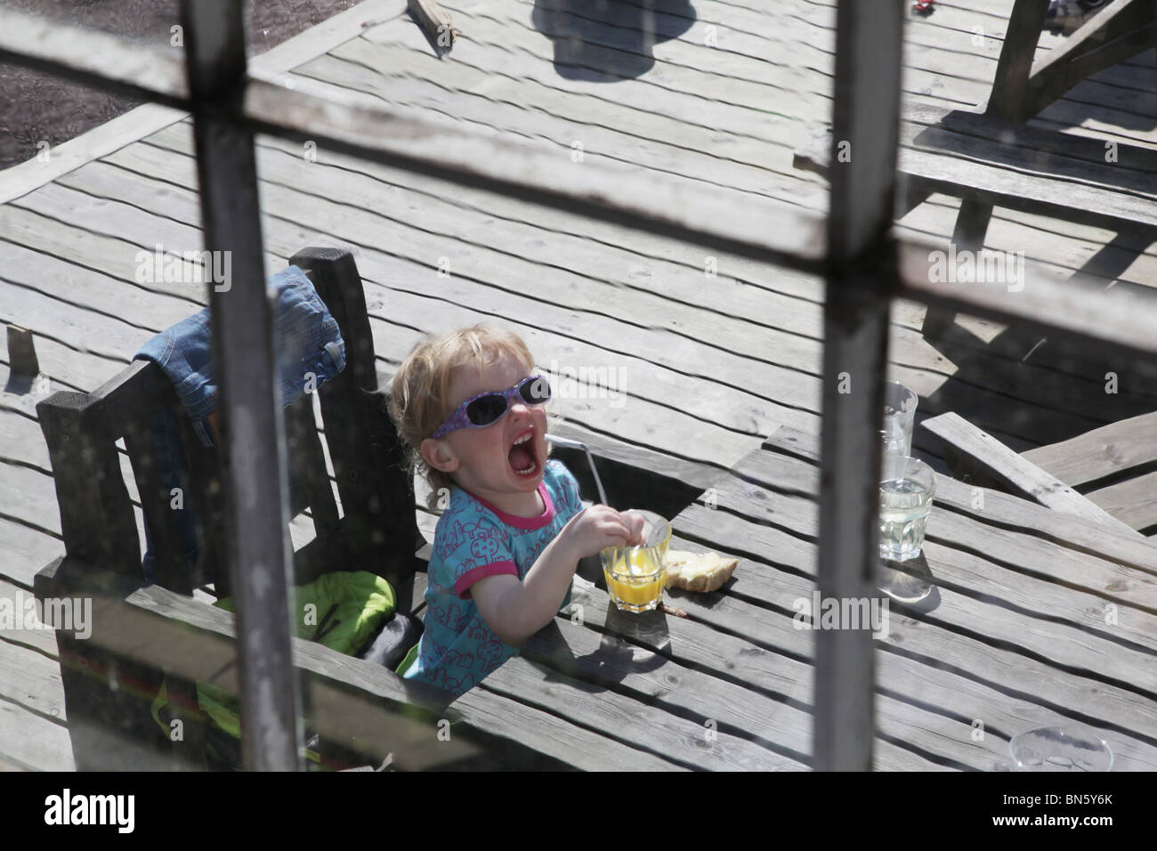 La bambina fotografata attraverso una bevanda alla finestra mentre beveva succo d'arancia sedeva all'aperto a prendere un volto che si divertiva con il MODELLO RILASCIATO Foto Stock