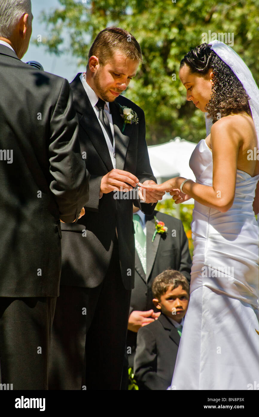 Caucasian lo sposo mette l'anello nuziale sul dito del suo mixed-gara sposa a loro formale all'aperto la cerimonia nuziale. Modello di rilascio Foto Stock