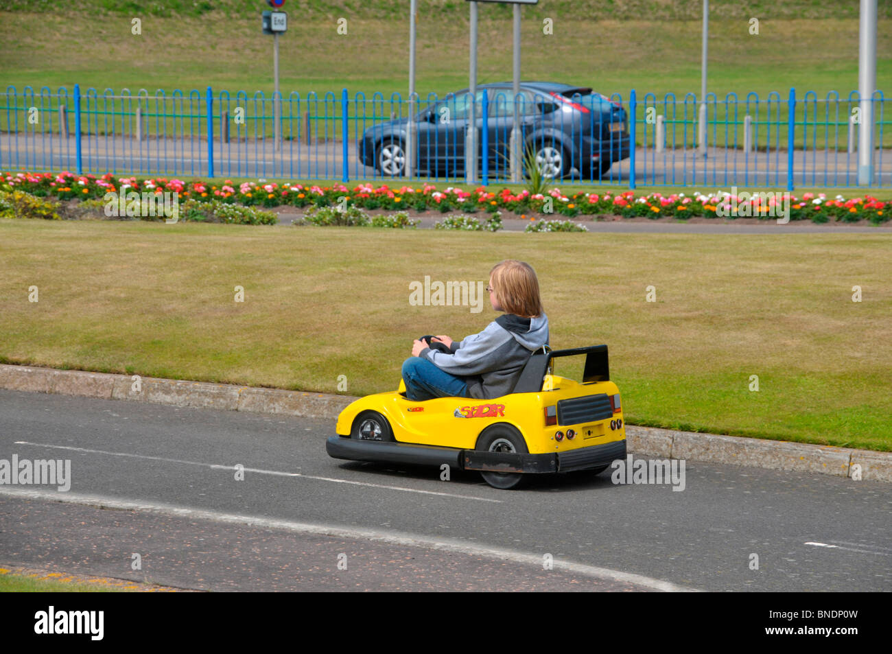 Un ragazzo giovane pilota un auto in miniatura - go-cart - attorno a un set di portate con una vera e propria auto sulla strada principale dietro di lui. Foto Stock