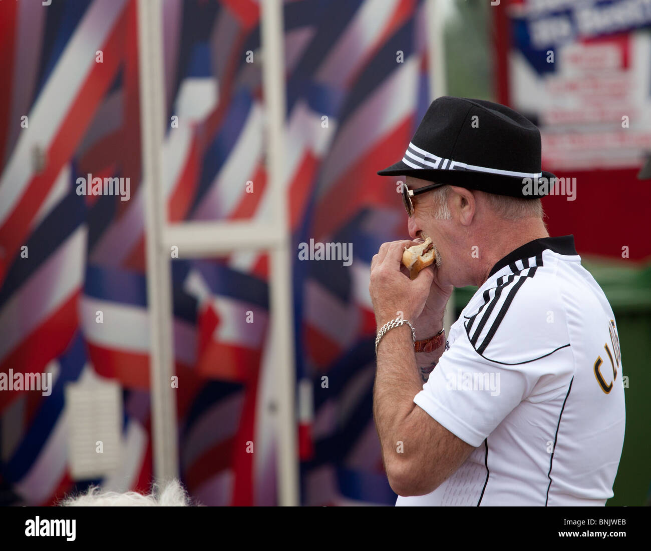 Un uomo che indossa una camicia dei tifosi di calcio prende un grosso boccone da UN hamburger mentre si allontana da un fast food in un evento all'aperto Fairford, regno unito Foto Stock