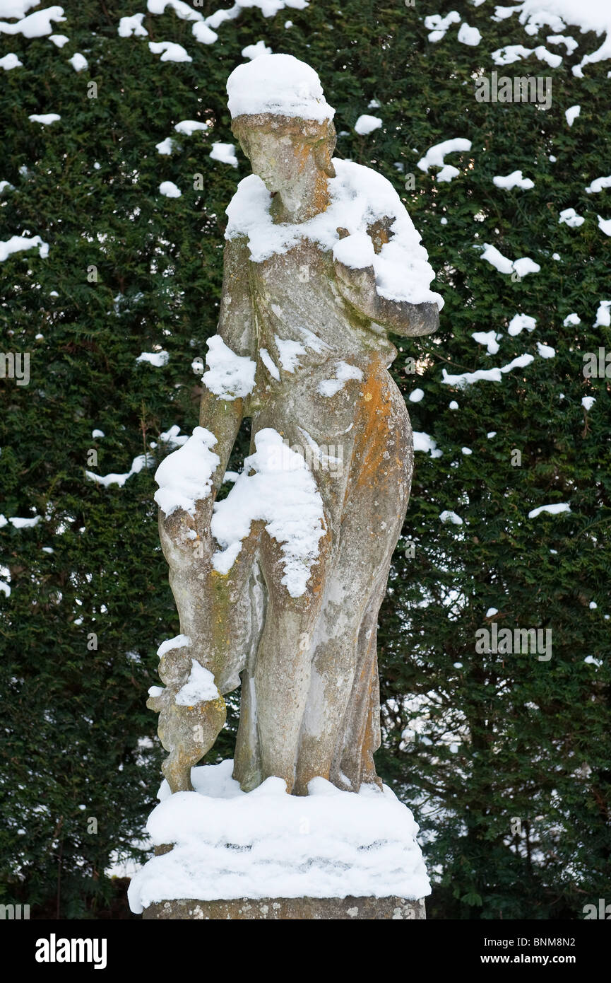Statua innevata di una ragazza con il suo cane da compagnia di fronte a una siepe di tasso in un giardino inglese in inverno Foto Stock