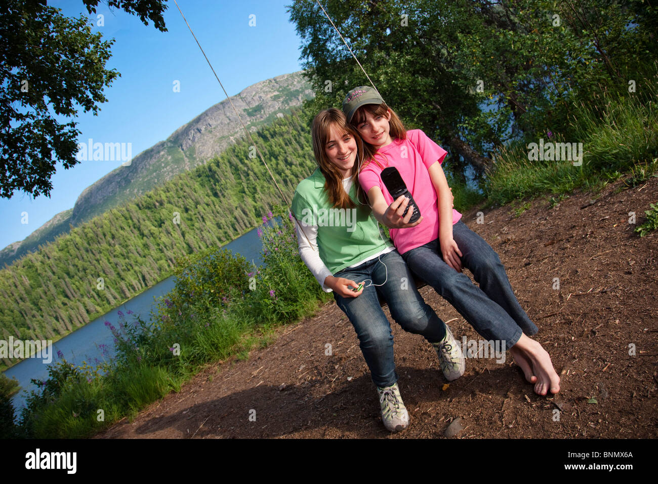 Due giovani ragazze seduto su una corda swing guardando le loro foto sul telefono cellulare, Byers Lago, Denali State Park, Alaska Foto Stock
