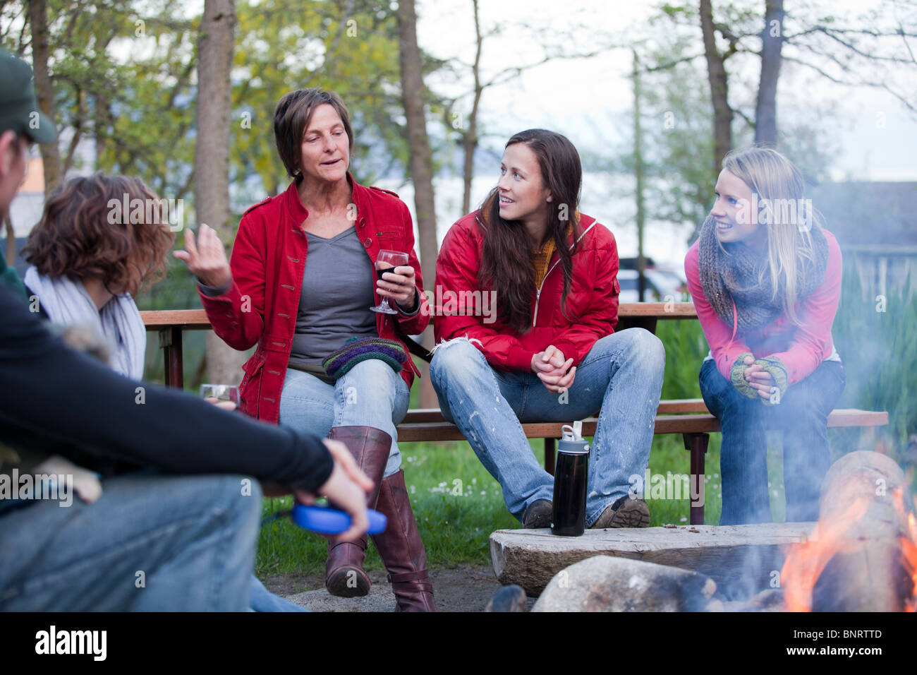Un gruppo di amici sedersi e gustare le bevande mentre si parla attorno a un fuoco di campo. Foto Stock
