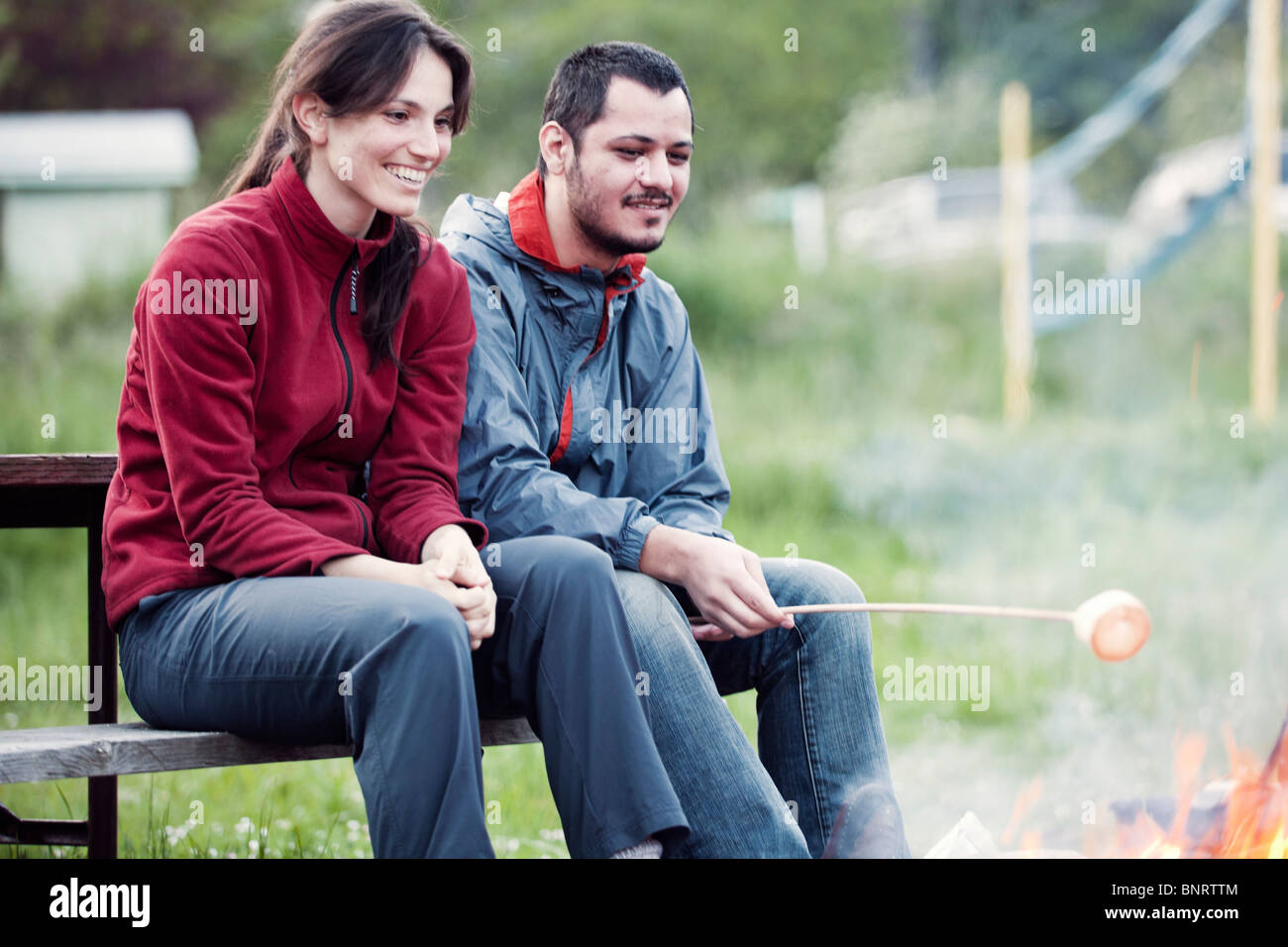 Una giovane coppia di sedersi su un banco di picnic e arrostire un marshmallow in corrispondenza di un fuoco di campo. Foto Stock