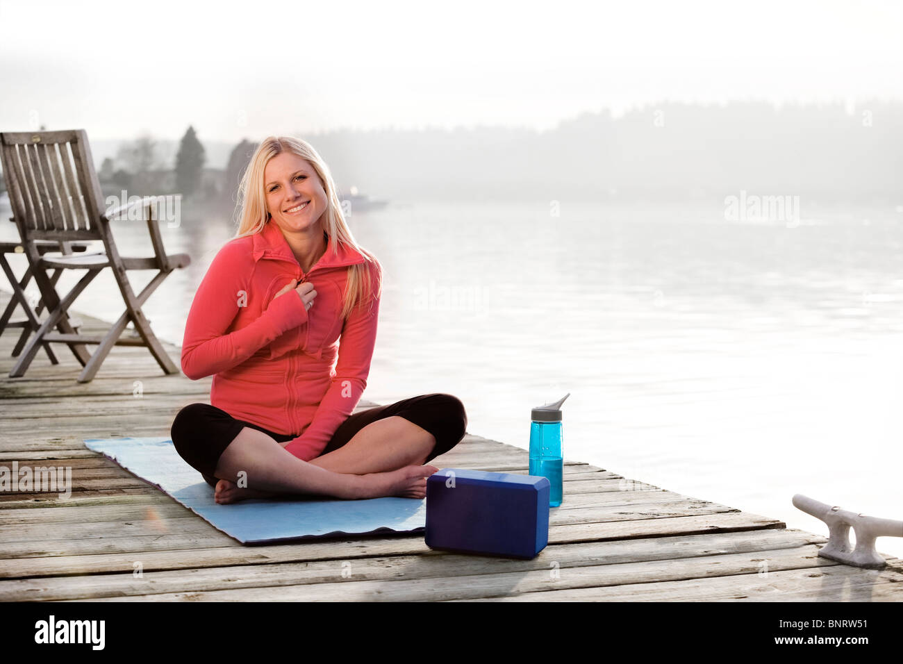 Una giovane donna appendere fuori su una dock sul lago Washington e sorridente alla fotocamera mentre è seduto su un materassino yoga. Foto Stock