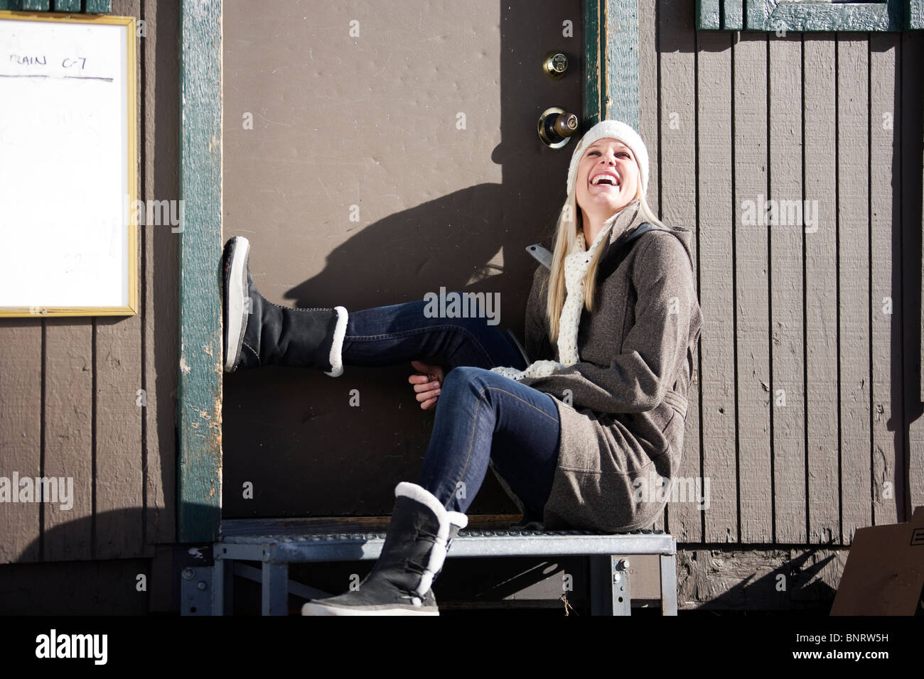 Una giovane donna vestita per il freddo si siede sui gradini di casa e risate. Foto Stock