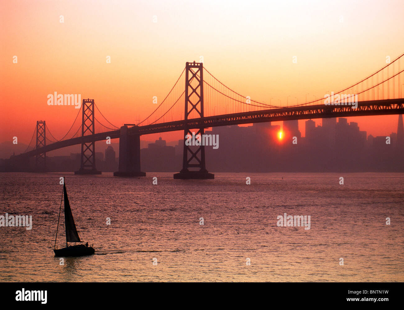 Barca a vela passando Oakland Bay Bridge sulla Baia di San Francisco con il tramonto spiata attraverso il profilarsi dello skyline della città Foto Stock