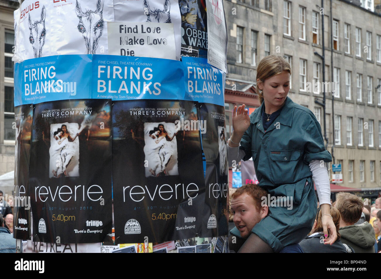 Una ragazza siede su un uomo di spalle di appendere manifesti sul Royal Mile durante la Edinburgh Fringe Festival. Foto Stock