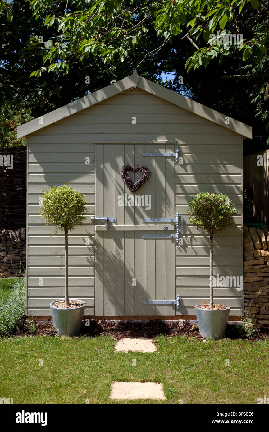 Grazioso giardino in legno tool shed con baia alberi standard su entrambi i lati della porta con il cuore a forma di corona sulla porta Foto Stock