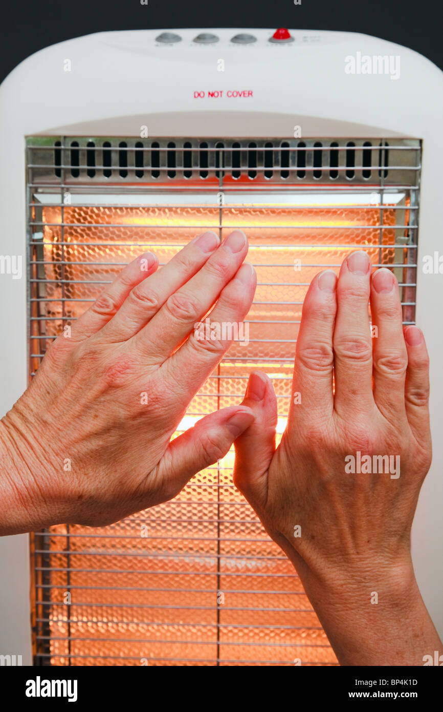 Scena quotidiana di una donna senior riscaldando le mani di fronte a bassa energia alogeno riscaldamento in camera. Inghilterra Regno Unito Gran Bretagna Foto Stock