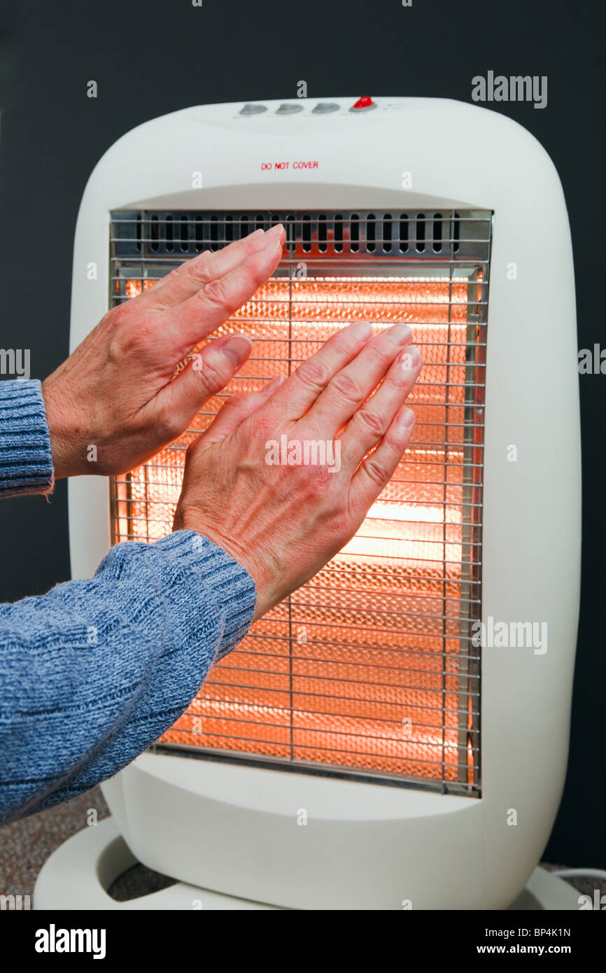 Un senior donna cercando di tenere in caldo riscaldando le mani di fronte a bassa energia alogeno riscaldatore camera d'inverno. Inghilterra Regno Unito Gran Bretagna Foto Stock