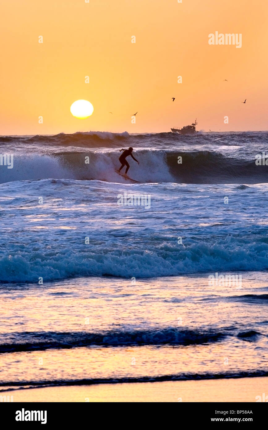 Un surfista in onde al tramonto con la silhouette di una barca in massa posteriore Foto Stock