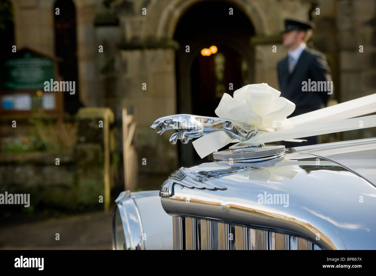 Primo piano di un'auto Jaguar decorata con nastri bianchi per un matrimonio, con autista fuori fuoco in piedi fuori da una chiesa, in lontananza. Foto Stock