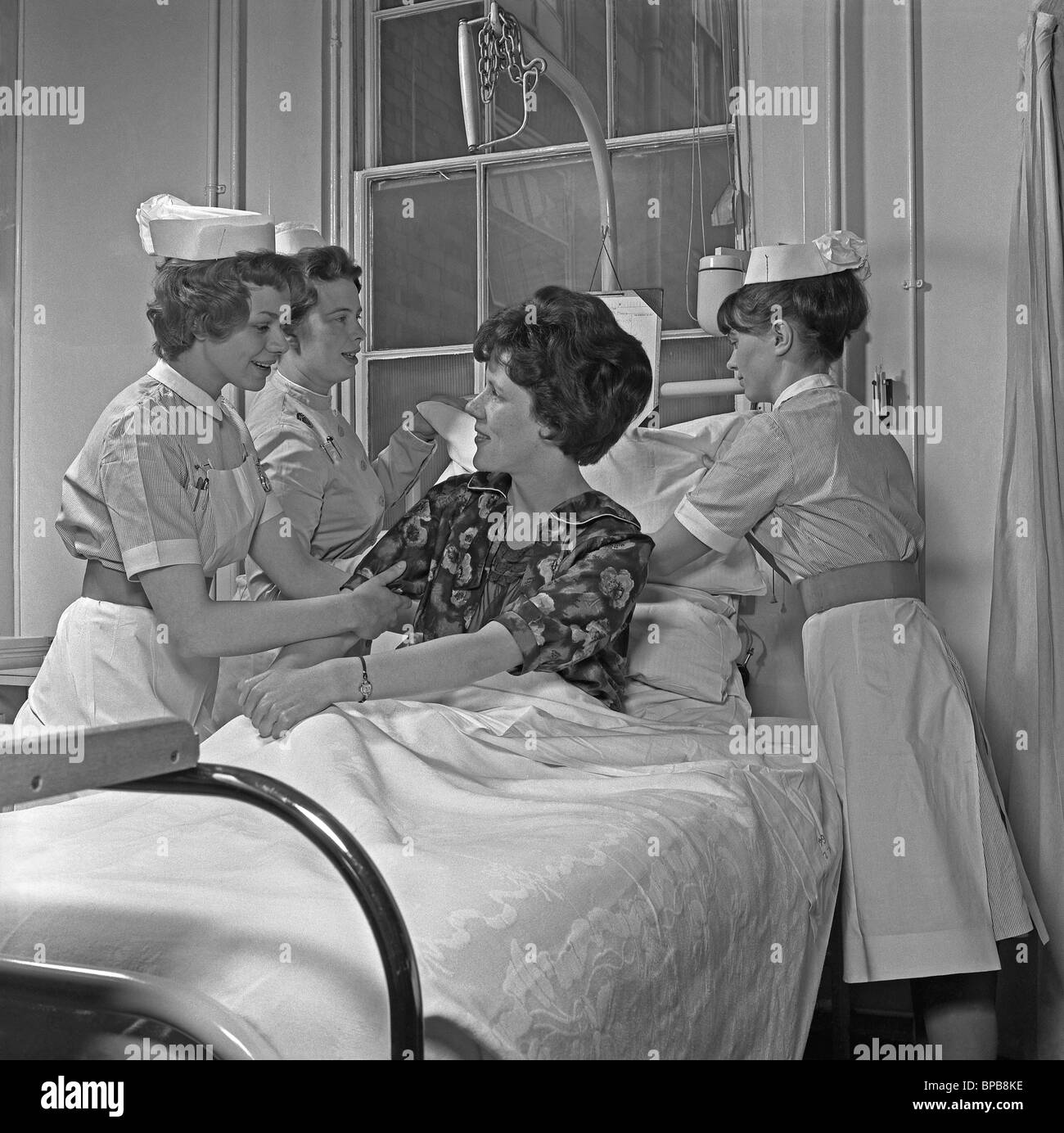 Ospedale infermieri riuniti intorno a un paziente nel letto la sua regolazione dei cuscini, c. 1960 Foto Stock