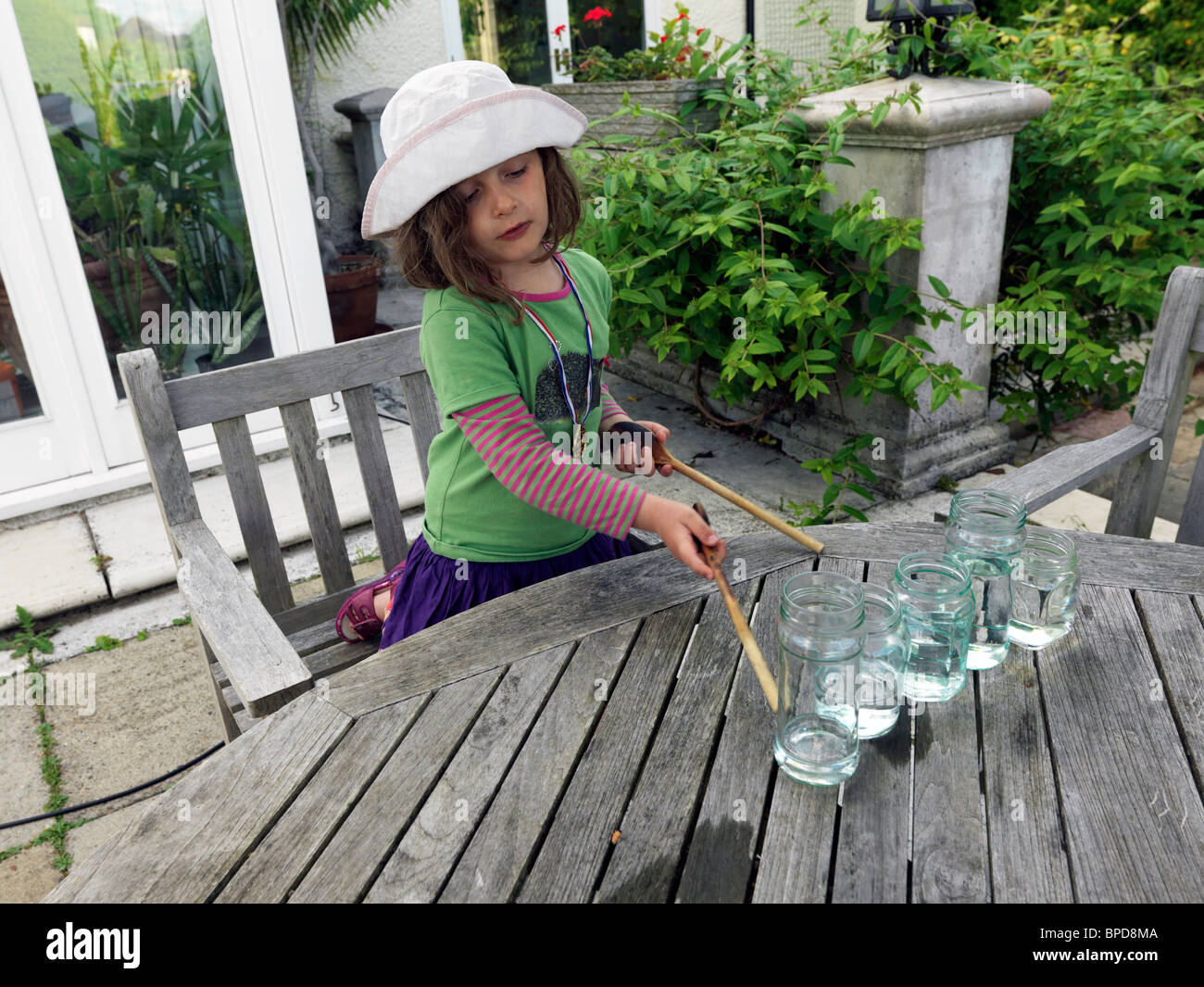 Ragazza giovane fare musica utilizzando vasetti di vetro pieno di acqua e li colpisce con cucchiai di legno Foto Stock