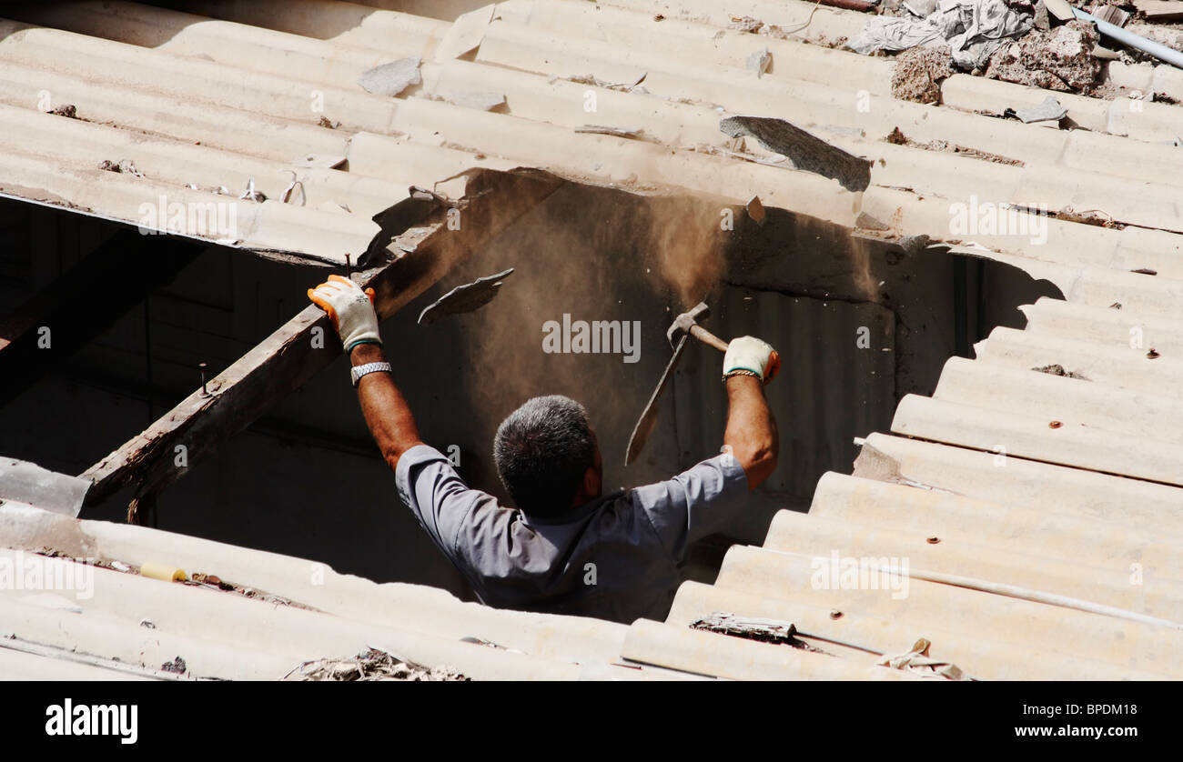 Uomo spagnolo rimuovendo il vecchio tetto in amianto con un martello. Nessuna maschera di respirazione o di abbigliamento protettivo in mezzo alle nuvole di polvere Foto Stock