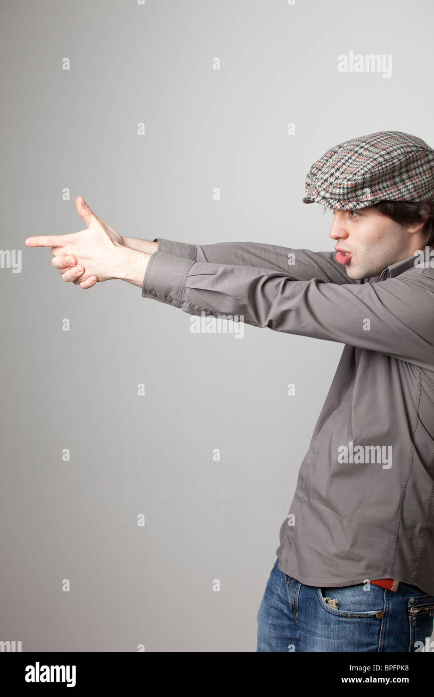 Giovane uomo con un cappello piatto e una camicia grigia, imitando una pistola con le mani, puntando a lato con un'espressione mirata. Foto Stock