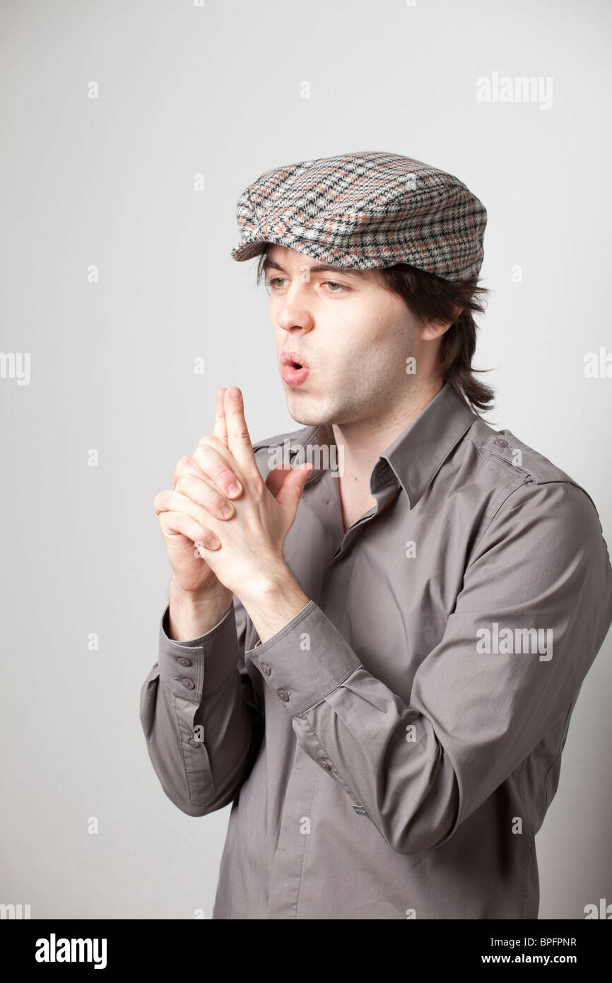 Giovane con una camicia grigia e un cappello piatto a quadri che fa un gesto di pistola con un'espressione sorpreso su uno sfondo neutro. Foto Stock