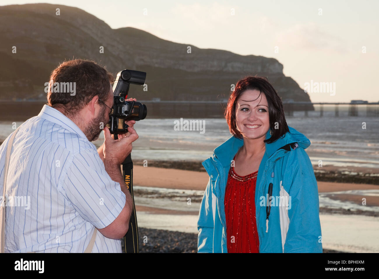 Fotografo a fotografare un giovane modello femminile sorridente sul lungomare utilizzando il flash della fotocamera. Il Galles, Regno Unito, Gran Bretagna Foto Stock