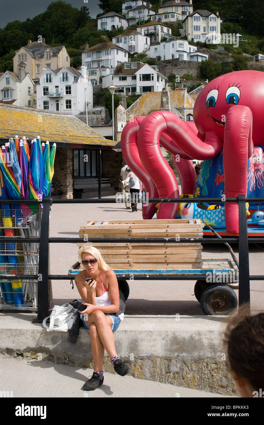 Donna seduta sulla banchina di sms sul suo telefono cellulare, con una rosa di polpo fairground ride in background. Looe, Cornwal Foto Stock