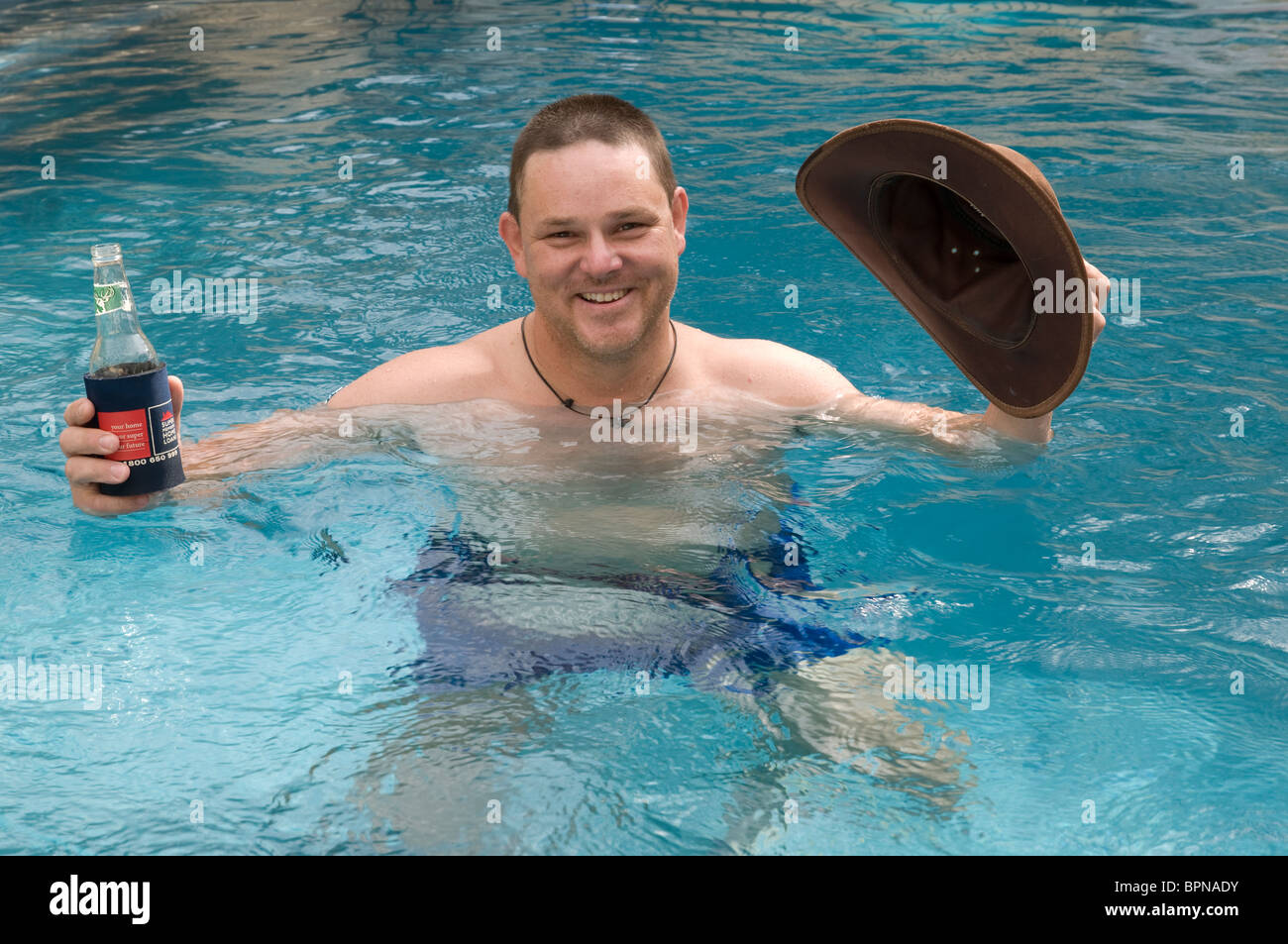 Uomo in una piscina tenendo un cappello e una bottiglia di bevanda Foto Stock