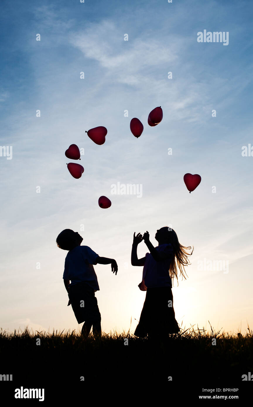 Silhouette di un giovane ragazzo e ragazza che gioca con la forma di cuore palloncini al tramonto Foto Stock