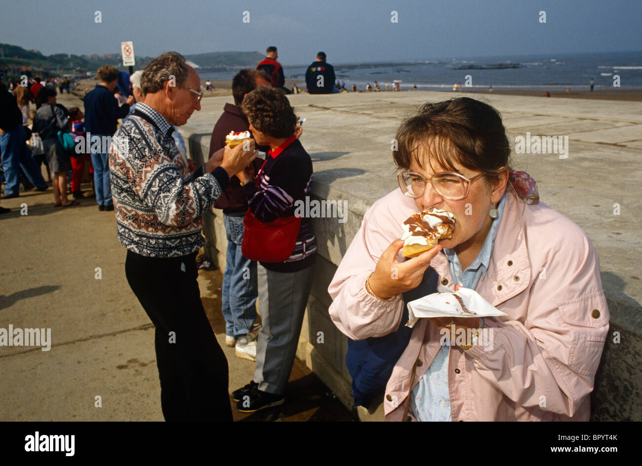 Vacanziere affonda i suoi denti in una crema di tortina farcita che trasuda su un tovagliolo mentre sul lungomare a Scarborough. Foto Stock